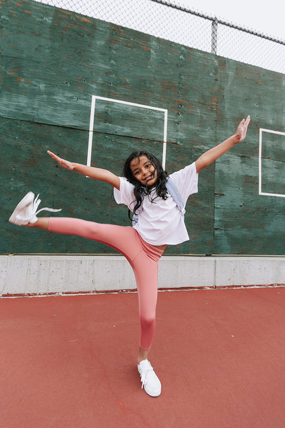 Young girl on a tennis court wearing Everyway kids activewear. Featuring a White Daily Tee and Dusty Rose All Day Leggings.