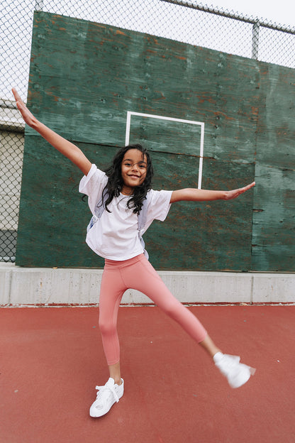 Young girl on a tennis court wearing Everyway kids activewear. Featuring a White Daily Tee and Dusty Rose All Day Leggings.